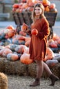 Portrait of happy woman with ripe orange pumpkins in hands near wagon with orange pumpkin on farmers market in brown sweater,