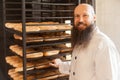 Portrait of happy young adult baker with long beard in white uniform standing in his workplace and carrying shelves with bread at Royalty Free Stock Photo