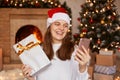 Portrait of happy young adult blogger woman wearing white sweater and santa claus hat, posing near fireplace and xmas tree, having Royalty Free Stock Photo