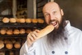 Portrait of happy young adult hungry baker with long beard in white uniform standing in his workplace and tasty fresh baked long Royalty Free Stock Photo