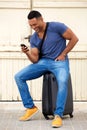 Happy young african man sitting on suitcase and using cell phone Royalty Free Stock Photo