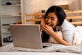 Portrait of happy young Asian girl in casual clothing lying down on bed while making a video call with laptop computer Royalty Free Stock Photo