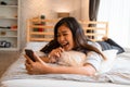 Portrait of happy young Asian girl in casual clothing lying down on bed while making a video call with smartphone in Royalty Free Stock Photo