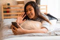 Portrait of happy young Asian girl in casual clothing lying down on bed while making a video call with smartphone and Royalty Free Stock Photo