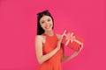 Portrait of a happy young asian woman holding gift box isolated over pink background. dressed in orange dress and sunglasses on Royalty Free Stock Photo