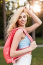 Portrait happy young blonde woman with cute smile in fashionable blue top with pink sports backpack in white skirt in forest at