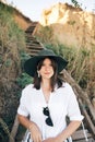 Portrait of happy young fashionable woman relaxing on tropical island. Stylish boho girl in hat sitting on wooden stairs at sandy Royalty Free Stock Photo