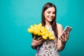 Portrait of a happy young girl in dress use mobile phone while holding big bouquet of yellow tulips isolated over color background Royalty Free Stock Photo