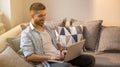 Portrait of happy and young handsome man, wearing casual clothes, working on a laptop and sitting on his sofa at home. Royalty Free Stock Photo