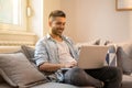 Portrait of happy and young handsome man, wearing casual clothes, working on a laptop and sitting on his sofa at home. Royalty Free Stock Photo