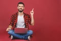 Portrait of a happy young man in casual holding laptop computer while sitting on a floor and pointing finger up isolated over red Royalty Free Stock Photo