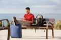 Happy young man sitting on bench with luggage and listening to music with mobile phone and earphones Royalty Free Stock Photo