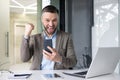 Portrait of a happy young man sitting at an office desk, holding a phone and showing a winning gesture of success with Royalty Free Stock Photo