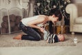 Portrait of happy young mother with child fooling around near Christmas tree and fireplace. Woman holds baby`s legs on