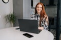 Portrait of happy young red-haired woman sitting at desk with laptop computer and mobile, smiling looking away. Royalty Free Stock Photo