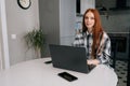 Portrait of happy young red-haired woman sitting at desk with laptop computer and mobile, smiling looking away. Royalty Free Stock Photo