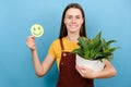 Portrait of happy young woman gardener holding funny face emoticon and potted plant, smiles broadly wears special uniform, Royalty Free Stock Photo