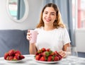 Portrait of a happy young woman holding a strawberry smoothie in her hand and ripe strawberries on table at home Royalty Free Stock Photo