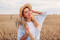 Portrait of happy young woman walking among wheat in summer field wearing straw hat holding bundle of ripe wheat