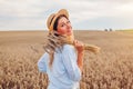 Portrait of happy young woman walking among wheat in summer field wearing straw hat holding bundle of ripe wheat