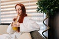 Portrait of happy young woman in white bathrobe drinking cocktail from straw with closed eyes sitting on sofa by window Royalty Free Stock Photo