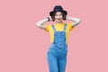 Portrait of happy young woman in yellow t-shirt, blue denim overalls with makeup and black hat standing posing with raised arms Royalty Free Stock Photo