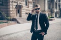 Portrait of a harsh agent outdoors in the city. He looks stunning and severe, wearing suit and sunglasses, fixing them and has