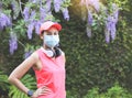 Asian woman in pink sportswear and medical face mask and headphones around neck,  standing hands on hips smiling and looking at Royalty Free Stock Photo