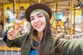 Portrait of a hippie girl in a hat who takes a selfie on a smartphone and shows the gesture of peace in the park near the attracti