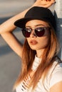 Portrait hipster modern stylish woman in  fashionable black cap in torn sexy T-shirt in fashion sunglasses relaxes on the street Royalty Free Stock Photo