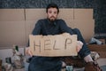 Portrait of homeless guy sitting on cardboard and holding a help cardboard in hands. He is looking straight at camera