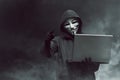 Portrait of hooded hacker with mask holding laptop while standing