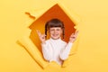 Portrait of hopeful little girl with braids wearing white turtleneck posing in torn hole of yellow paper wall, crossing fingers, Royalty Free Stock Photo