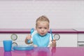 Portrait Of Hungry todler in a blue bib with fork and knife in his hands In High Chair in the modern kitchen ready to