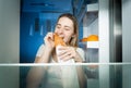 Portrait of young hungry woman standing at open refrigerator at night and eating bread Royalty Free Stock Photo