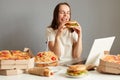Portrait of hungry woman watching movie laptop computer, eating hamburger while resting, having cheat meal isolated over gray