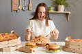 Portrait if amazed excited extremely hungry woman wearing white T-shirt sitting at table in kitchen, delivering lots jink food