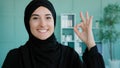 Portrait indoors Muslim Islamic girl smiles toothy wide at camera showing hand ok sign. Female Arabian student shows Royalty Free Stock Photo