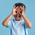 Portrait of inquisitive nosy little boy looking through fingers shaped like binoculars against a blue studio background