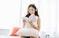 Portrait of inspired beautiful smiling Asian woman sitting on white bed reading book and listening to music in headphones.