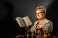 Portrait of an intelligent young woman in elegant glasses reading a book sitting in a chair in a dark room Royalty Free Stock Photo