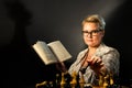 Portrait of an intelligent young woman in elegant glasses reading a book sitting in a chair in a dark room Royalty Free Stock Photo