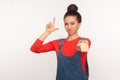 Portrait of irritated girl with hair bun in denim overalls showing loser gesture and pointing to camera, blaming for defeat Royalty Free Stock Photo