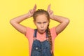 Portrait of irritated little girl in denim overalls showing bull horn gesture, looking at camera with hostile angry expression