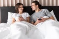 Portrait of a jelous young man looking at his girlfriends mobile phone while sitting together in bed at home