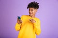 Portrait of joyful african american man wearing bluetooth earpod smiling and holding smartphone, isolated over violet background