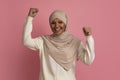 Portrait of joyful arabic woman in hijab celebrating success over poink background