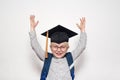 Portrait of a joyful blond boy in big glasses, academic hat and a backpack. Hands up. White background Royalty Free Stock Photo