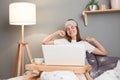 Portrait of joyful cheerful positive woman wearing white t-shirt and blindfold sitting in bed in front of laptop, being in good Royalty Free Stock Photo