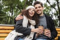 Portrait of joyful couple man and woman 20s drinking takeaway coffee from paper cups, while sitting on bench in green park Royalty Free Stock Photo